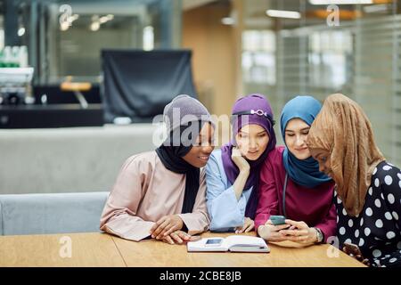 Beautiful young muslim multiethnic group of girls in long smart traditional dresses and hijabs looking at smart phone in hand of asian woman while stu Stock Photo