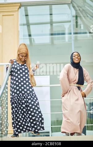 Couple of muslim African 20s girlfriends with hijab or islamic head scarfs and islamic traditional clothes having fun visiting modern glass shopping m Stock Photo