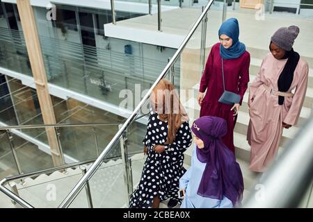 Multicultural group of female Arabian students, dressed in national dresses and scarfs going down the stairs of university , sharing ideas with each o Stock Photo