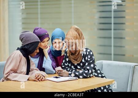 Beautiful young muslim multiethnic group of girls in long smart traditional dresses and hijabs looking at smart phone in hand of african woman while s Stock Photo