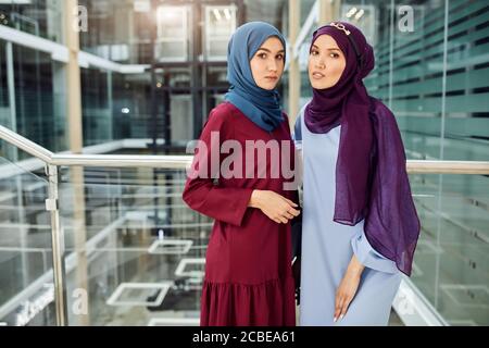 Portrait of two islamic young women dressed in muslim wear with hijabs on head looking at camera while standing against glass and steel loft interior Stock Photo