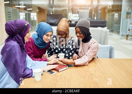 Portrait of smart elegantly dressed caucasian and african muslim female friends looking at hands of one of women admiring manicure while sitting in ci Stock Photo