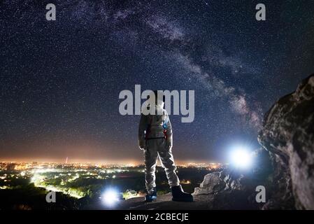 Mission specialist astronaut standing on rocky hill and looking at beautiful starry sky with milky way. Space traveler wearing white space suit and helmet. Concept of human space exploration. Stock Photo