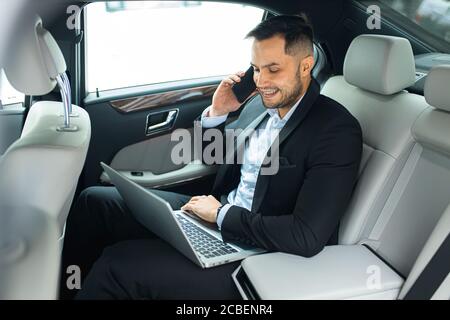 Handsome bearded man working on laptop while talking on phone in his luxurious car, side view Stock Photo