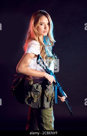 Beautiful caucasian female private military contractor with long blonde hair, dressed in tactical uniform, armed with riffle posing isolated in studio Stock Photo