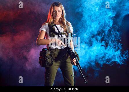 Woman soldier in military camouflage uniform protected with headphones, body armour, holding assault rifle standing against dark smoky background Stock Photo