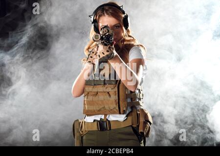 Airsoft female shooter playing strikeball, sets her sights on enemy with automatic assault rifle. Studio shot, dark smoky background. Stock Photo