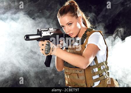 Airsoft female shooter playing strikeball, sets her sights on enemy with automatic assault rifle. Studio shot, dark smoky background. Stock Photo