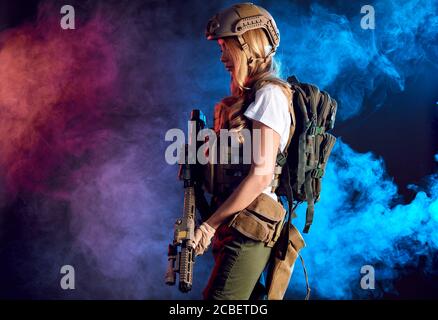 Heavily armed and well-equipped female soldier in battle helmet holding assault rifle isolated on dark smoky battlefield. Paint ball and laser tag spo Stock Photo