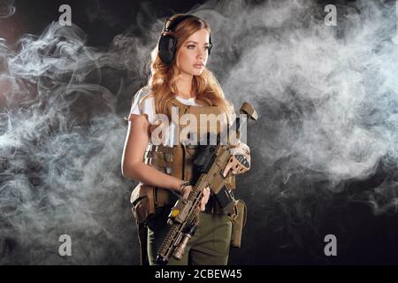 Woman soldier in military camouflage uniform protected with headphones, body armour, holding assault rifle standing against dark smoky background Stock Photo