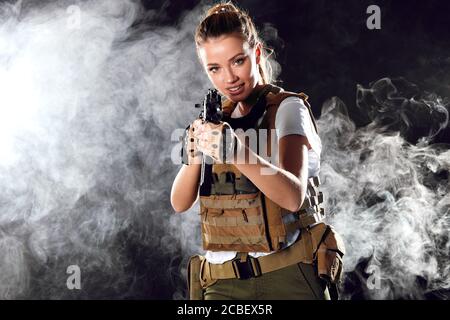 Airsoft female shooter playing strikeball, sets her sights on enemy with automatic assault rifle. Studio shot, dark smoky background. Stock Photo
