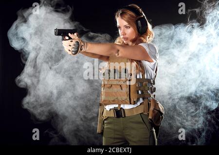 Woman in protective armour and headphones holds gun in hands, aiming over dark smoky background. Army, dedication, weapon, technology. Stock Photo