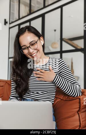 Attractive smiling young asian business woman relaxing on a leather couch at home, during video call on laptop computer, wearing earphones