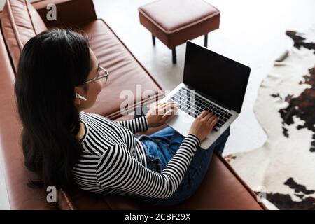 Top view of an attractive smiling young asian business woman relaxing on a leather couch at home, working on laptop computer Stock Photo
