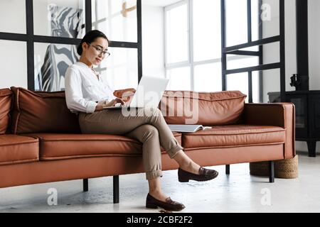 Attractive smiling young asian business woman relaxing on a leather couch at home, working on laptop computer Stock Photo