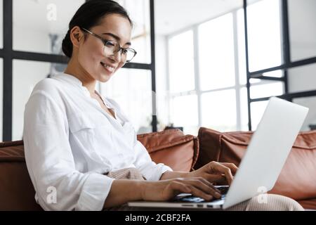 Attractive smiling young asian business woman relaxing on a leather couch at home, working on laptop computer Stock Photo