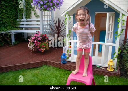 Four year old, white girl, stood on a pink, plastic, child’s chair screaming in front of a blue and white Wendy house. England UK Stock Photo