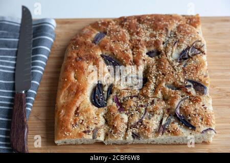 A large piece of freshly baked foccacia topped with red onion slices and sesame seeds on a chopping board Stock Photo