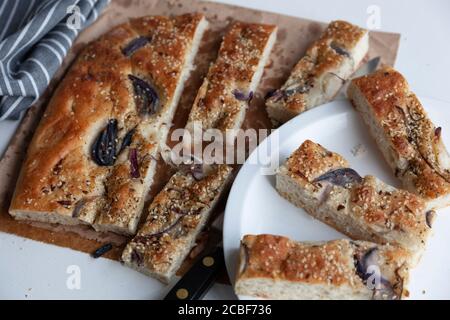 A large piece of freshly baked and sliced foccacia topped with red onion and sesame seeds on a board and plate Stock Photo