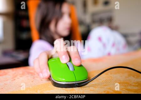 In the foreground is a child's hand on a computer mouse. In the background, a girl is sitting on a chair. Blurred background, colorful photo. Stock Photo