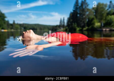 Beautiful pregnant young woman in a red dress floating arms wide in the water of a lake. Sunny day, warm Canadian summer. Spiritual atmosphere. Stock Photo