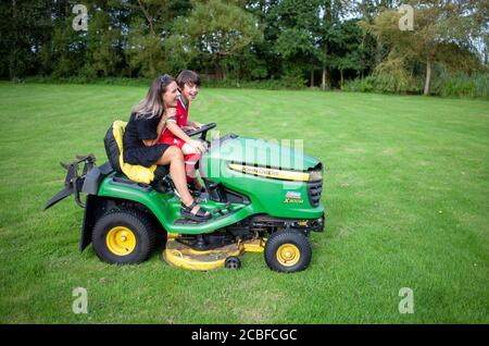 Mother and son riding a green and yellow John Deere tractor mower in a large English garden, Lancashire, England, UK Stock Photo