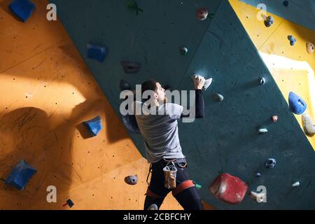 Rare view of unrecognizable male rock-climber with physical disability training at colourful artificial rock wall, hanging on two holds, has strong bi Stock Photo