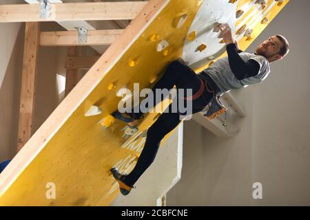 Handsome athletic male mountaineer with physical disability climbing at steep rock wall in indoor bouldering gym, wearing harness and chalk bag on wai