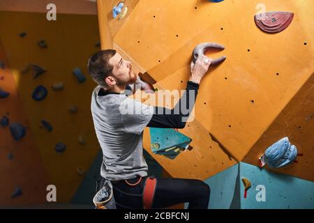 Side shot of well equipped sportsman training in colorful bouldering gym, climbing at difficult rock wall, overcomes his physical disability, has stro