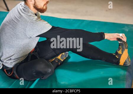Close up cropped shot of unrecognizable flexible boulderer doing stretching exercises before active climbing training, sitting at blue mat, dressed in Stock Photo