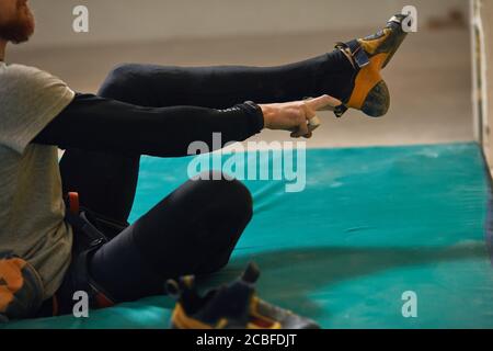 Close up view on strong legs, unrecognizable male climber with beard puts on climbing shoes, getting ready to intensive bouldering training, sitting o Stock Photo