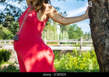 Close up and profile view of a pregnant woman in a sleeveless red dress standing barefooted under a tree, and holding with a hand to the trunk. Stock Photo