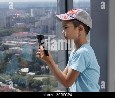 Yekaterinburg, Russia - July 18, 2019: A Muslim boy uses a smartphone against background of a panoramic window. Blurred view of city of Yekaterinburg Stock Photo