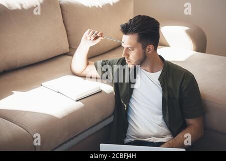 Thoughtful caucasian man reading something in the notebook and holding a laptop on the floor near the sofa Stock Photo