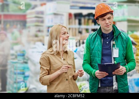 caucasian beautiful woman in market, warehouse worker consult her, introduce the assortment in store. worker in uniform and helmet explain, surprised Stock Photo