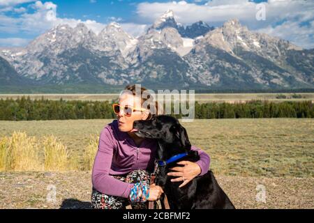 Woman hugs and holds her black labrador retriever dog in front of the Grand Teton National Park mountains in Jackson Wyoming as the dog licks her face Stock Photo
