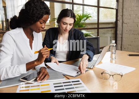 Two attractive smart multiethnic women entrepreneurs working over new project in a modern office, sitting at the desk Stock Photo