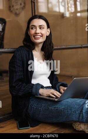 Attractive smiling young woman sitting at the office, leaning on a glass wall, using laptop computer Stock Photo