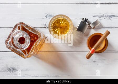 View of a crystal glass and decanter full of golden whisky, and cigar and petrol lighter, shot from above on a distressed white wooden background Stock Photo