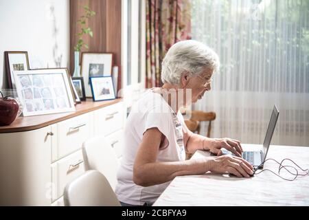 An elder lady with glasses staring at laptop's monitor while clicking ...