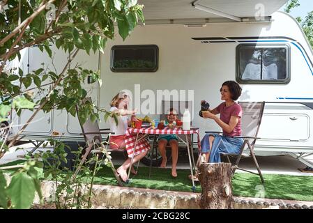 Single mother having breakfast with her two children in front of her caravan. They are enjoying their vacation. Stock Photo