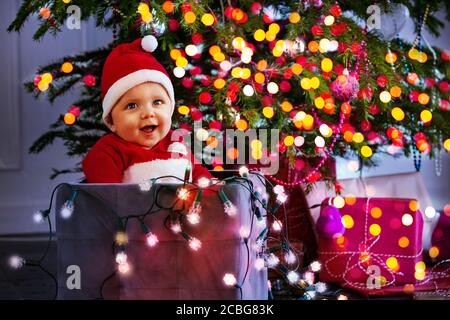 Little cute baby boy in Santa Claus hat holding illuminated lamps sitting in box under Christmas tree with blurred lights Stock Photo