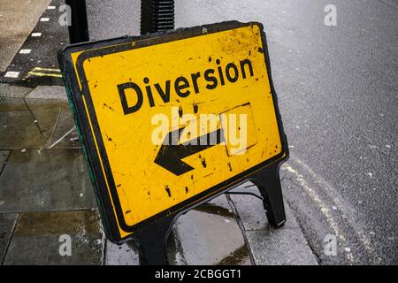 Yellow diversion sign with arrow pointing left on wet asphalt road. Stock Photo