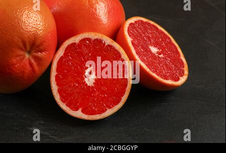 Pink grapefruit cut to half, more citrus fruits in background, on black board. Stock Photo