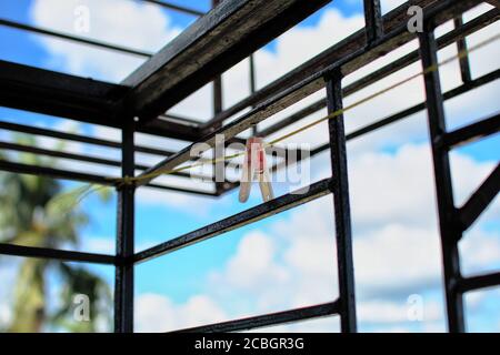 Isolated old faded pink plastic clothes peg hanging on yellow nylon rope inside railing balcony with blurred blue sky background Stock Photo
