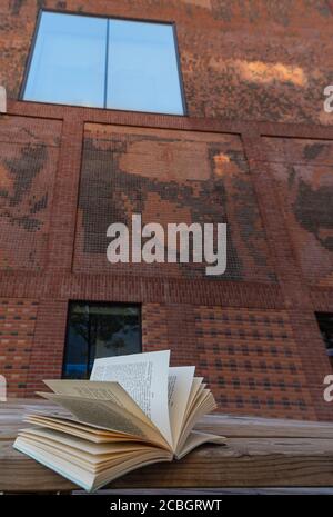 An open book resting on a bench in front of a red brick building with a stained glass window reflecting the blue sky Stock Photo