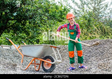 game of builder and construction. Outdoor activity concept. girl who is transporting rubble in a wheelbarrow. construction work. girl builder takes a shovel of rubble. hardworking youth. Stock Photo