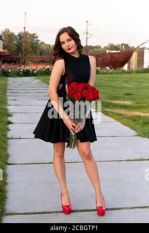 Attractive young birthday girl with brown hair in little black dress posing, smiling and holding a bouquet of red roses on background of wooden boat a Stock Photo