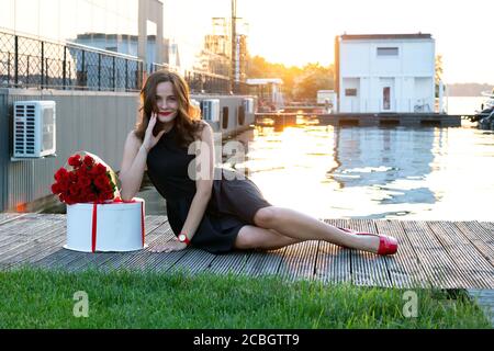 Attractive birthday girl with brown hair in little black dress sitting on the wooden footpath at sunset. Round box with birthday cake wrapped with red Stock Photo