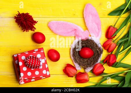 Tulips in pink or red colors on yellow wooden background. Happy holiday concept. Spring holiday composition. Bunch of flowers near Easter eggs in nest with bunny ears placed by present boxes. Stock Photo